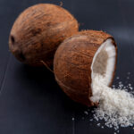 Close-up of desiccated coconut on a white background, displaying fine, dry flakes of coconut meat with a light beige color. The flakes are small and appear to be grated or shredded, commonly used as a topping or ingredient in various culinary dishes.