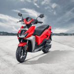 The scooter features a modern and minimalist design with a clean silhouette, sharp lines, and LED lighting. It has a compact body, with a digital instrument cluster, disc brakes, and alloy wheels. The scooter is powered by an electric motor and has a removable Lithium-Ion battery, offering a range of up to 75 km per charge. It also has regenerative braking for enhanced energy efficiency. The alt text highlights the scooter's contemporary design, electric powertrain, and eco-friendly features.